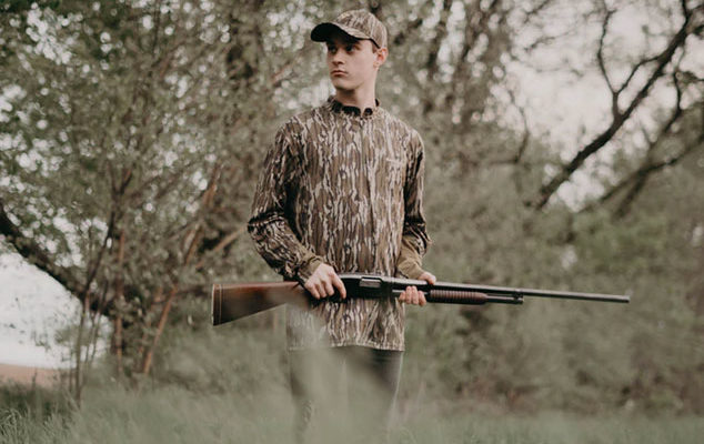 A hunter wearing camouflage stands with his rifle in the brush