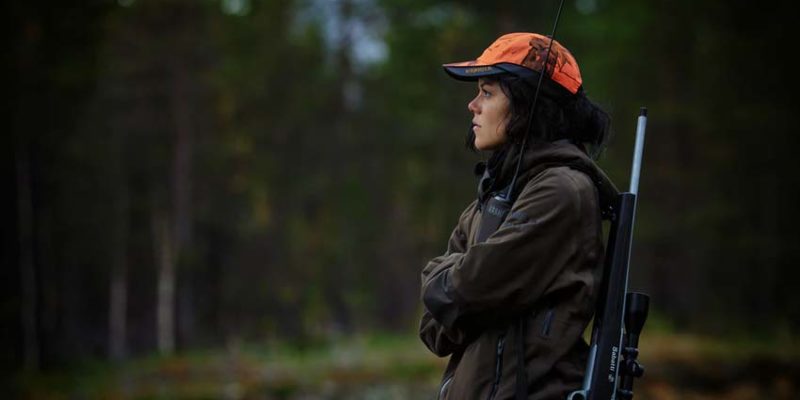 A hunter wearing an orange cap standing in the woods, with a rifle strapped to her back.