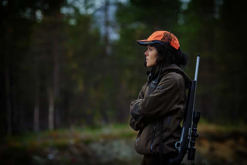 A hunter wearing an orange cap standing in the woods, with a rifle strapped to her back.