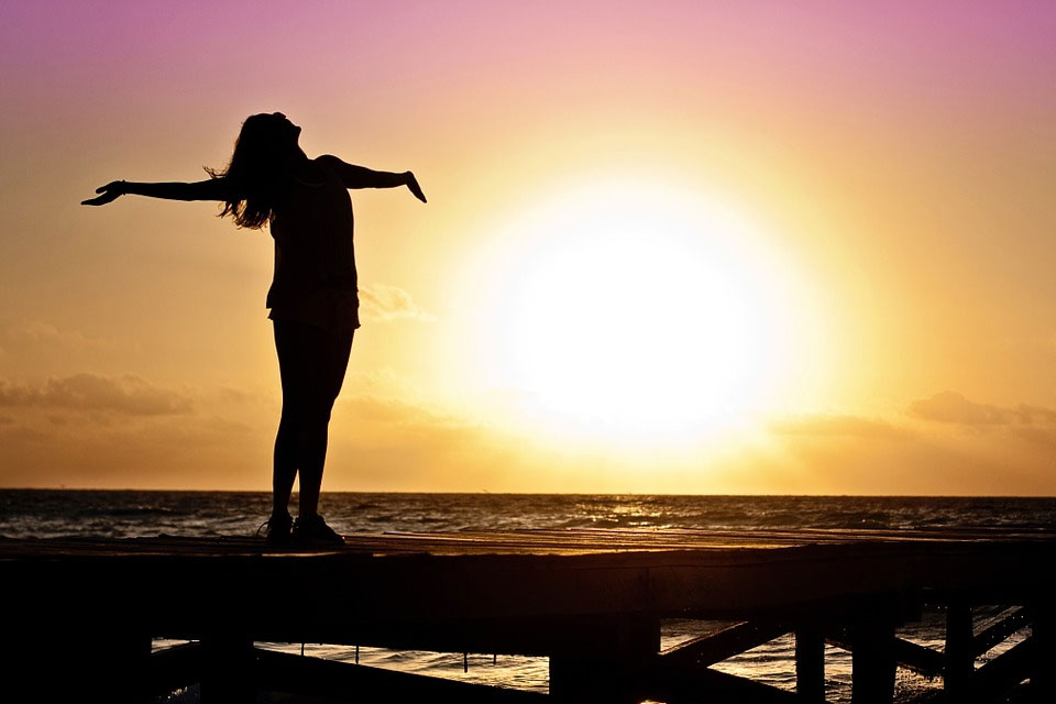 A woman lifts her arms and face to the sky in front of the sea at sunset