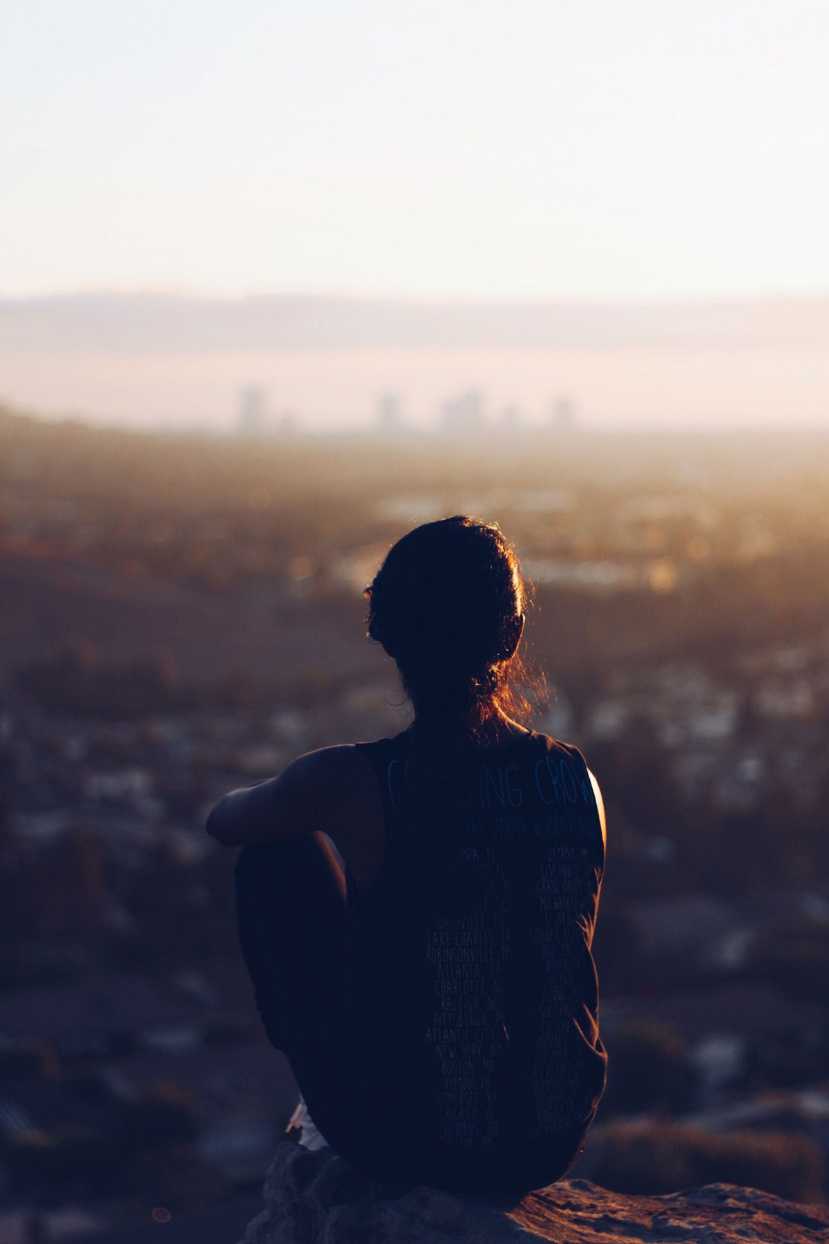 A woman in silhouette sitting on a rock, staring at a cityscape in the distance.