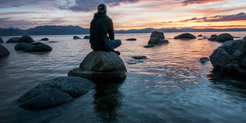 A man sitting alone on rocks in a lake at twilight
