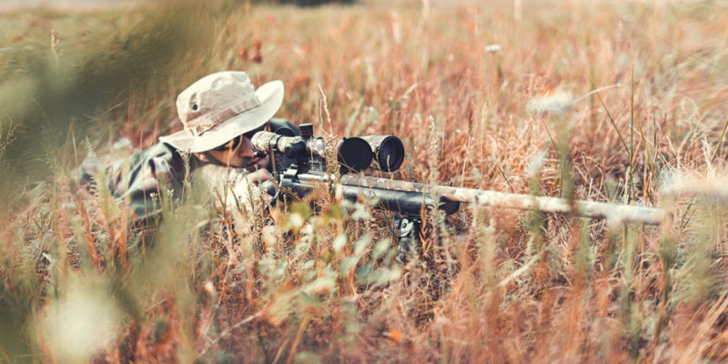 A man in camouflage lies in the grass with his hunting rifle, which is also camouflaged.