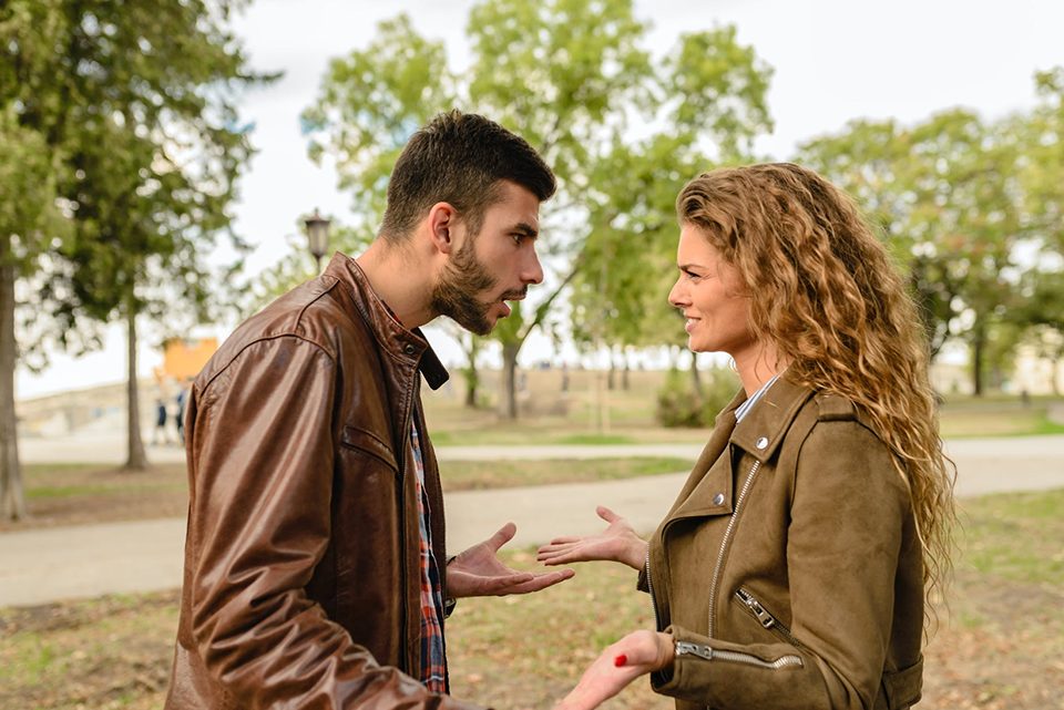 A man and a woman in brown leather jackets argue in a park.