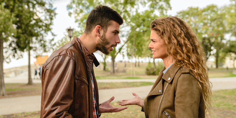 A man and a woman in brown leather jackets argue in a park.