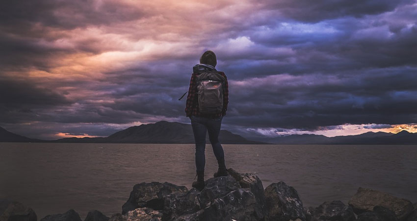 A backpacker stands on some rocks overlooking the sea and a distant shore.