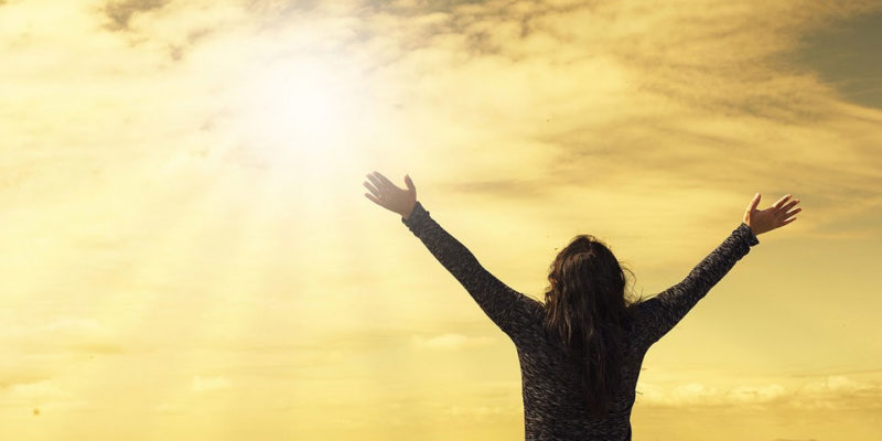 A woman on mountain ledge lifts arms up and stares up into the sky